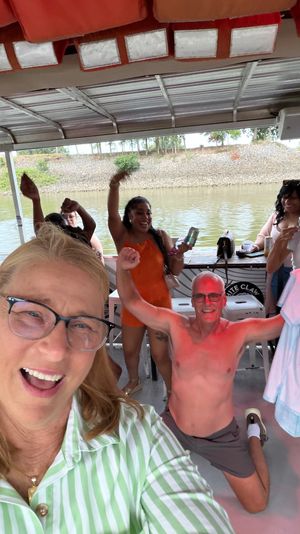 Cheerful group partying on a covered riverboat deck — selfie-style woman in the foreground, guests cheering with drinks, shirtless man posing, river shoreline and trees visible in the background.