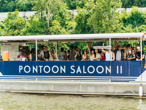 Energetic summer scene of a crowded blue pontoon party boat cruising a river, people cheering with drinks against a lush green shoreline