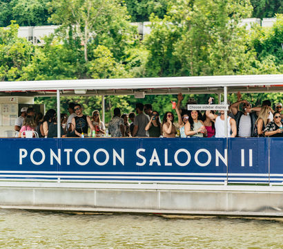 Energetic summer scene of a crowded blue pontoon party boat cruising a river, people cheering with drinks against a lush green shoreline