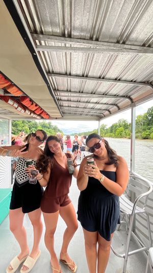 Three friends in sunglasses holding canned drinks and smiling on a covered pontoon boat during a sunny summer river cruise with a tree-lined shoreline in the background.