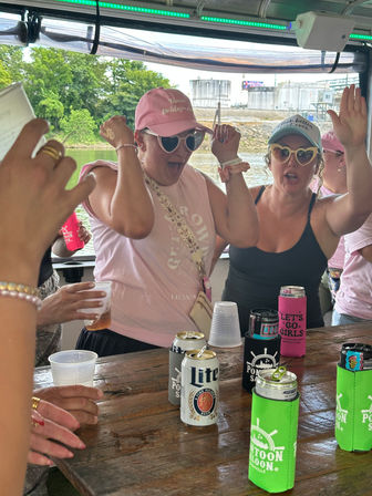Bachelorette crew partying on a pontoon boat river cruise — women in pink caps and heart-shaped sunglasses cheering over drinks and colorful koozies on a wooden table.