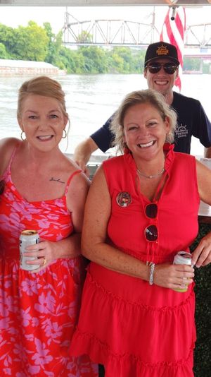 Group of six adults in red outfits smiling and holding drinks on a riverboat cruise, with green riverbank and a steel bridge visible in the background.