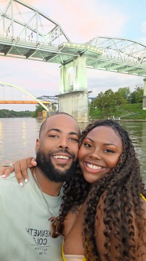 Smiling couple selfie on a riverboat at dusk with an illuminated steel truss bridge and arched bridge spanning the water