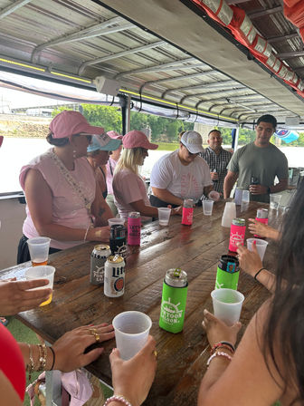 Cheerful group of people on a covered pontoon boat cruising a river, gathered around a wooden table with drinks in colorful koozies; several wearing pink shirts and caps on a sunny outing.
