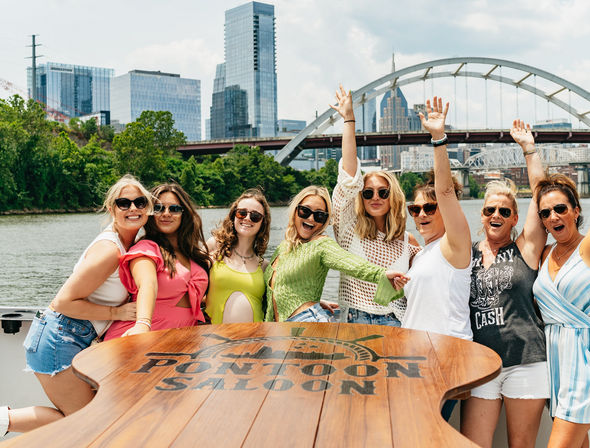 Eight friends in sunglasses partying on a pontoon boat — wooden branded table in foreground, river with green shoreline, arched bridge and modern city skyline under a sunny sky.