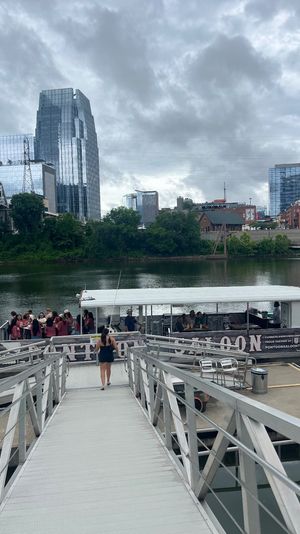 Dockside riverboat with passengers and American flag at a riverside gangway, framed by an urban downtown skyline of reflective glass towers under a cloudy sky.