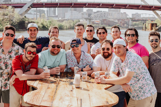 Group of friends at an urban riverfront under a city bridge and skyline, leaning on a rustic wooden table and holding beer cans in casual summer shirts and sunglasses.