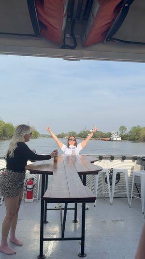 Two women on a riverboat deck by a long wooden high-top table, one cheering with arms raised, life jackets overhead and a tugboat passing on a tree-lined river under a clear blue sky.