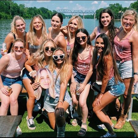 Group of young women posing for a cheerful summer riverfront photo, wearing casual tanks and denim shorts, holding canned drinks and a large face cutout, with a steel bridge over the water in the background.