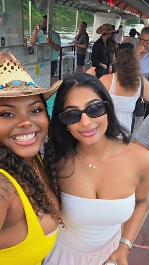 Two friends smiling selfie on a crowded river cruise boat — one in a straw cowboy hat and yellow dress, the other wearing sunglasses and a white tube top, summer party vibe.