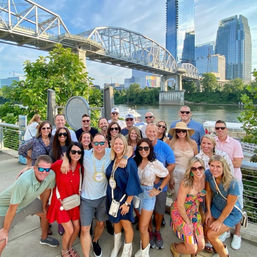Cheerful group of adults posing on a riverfront promenade beneath a steel truss bridge, with downtown glass skyscrapers, trees and a calm river in the background.