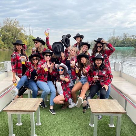 Lively boat party on a river: a group in red plaid shirts and black cowboy hats cheering with drinks on a boat deck, trees and cloudy sky in the background