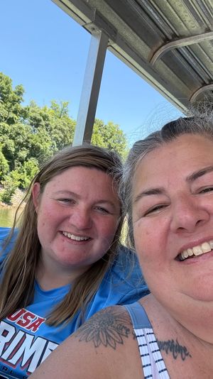 Two smiling women taking a sunny selfie on a covered boat over a calm lake with green trees and blue sky in the background.