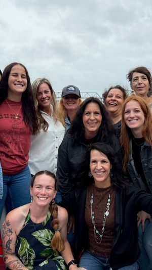 Smiling group of women aboard a river boat cruise, posing by a wooden table with a stone tower, metal bridge and tree-lined riverbank under an overcast sky.