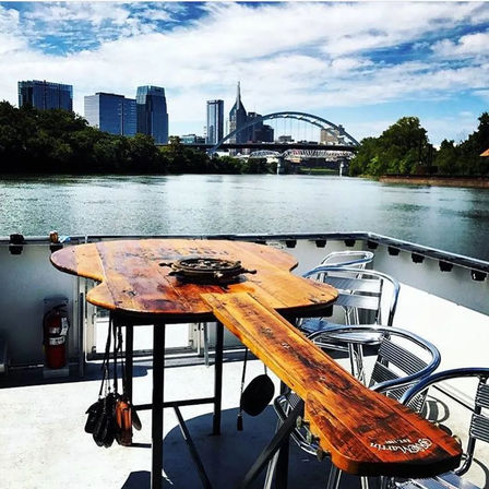 Paddle-shaped wooden table and metal chairs on a riverboat deck, looking across the Cumberland River toward downtown Nashville skyline and a graceful arched bridge under a blue sky