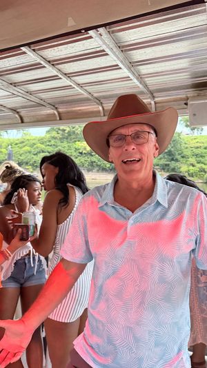 Smiling man in a cowboy hat and tropical shirt at a lively riverboat party, surrounded by friends chatting near a green riverbank