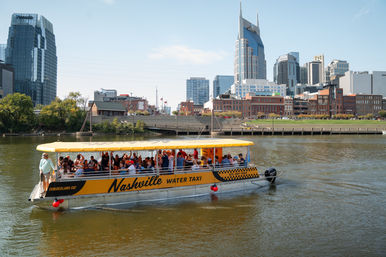 Yellow water taxi filled with passengers cruising the Cumberland River with downtown Nashville skyline and a distinctive twin‑spired skyscraper rising behind brick riverfront buildings