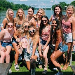 Smiling group of women posing on a sunny riverfront dock with a steel bridge in the background, wearing summer tanks and denim shorts, holding canned drinks and an oversized face cutout — lively summer river party scene.