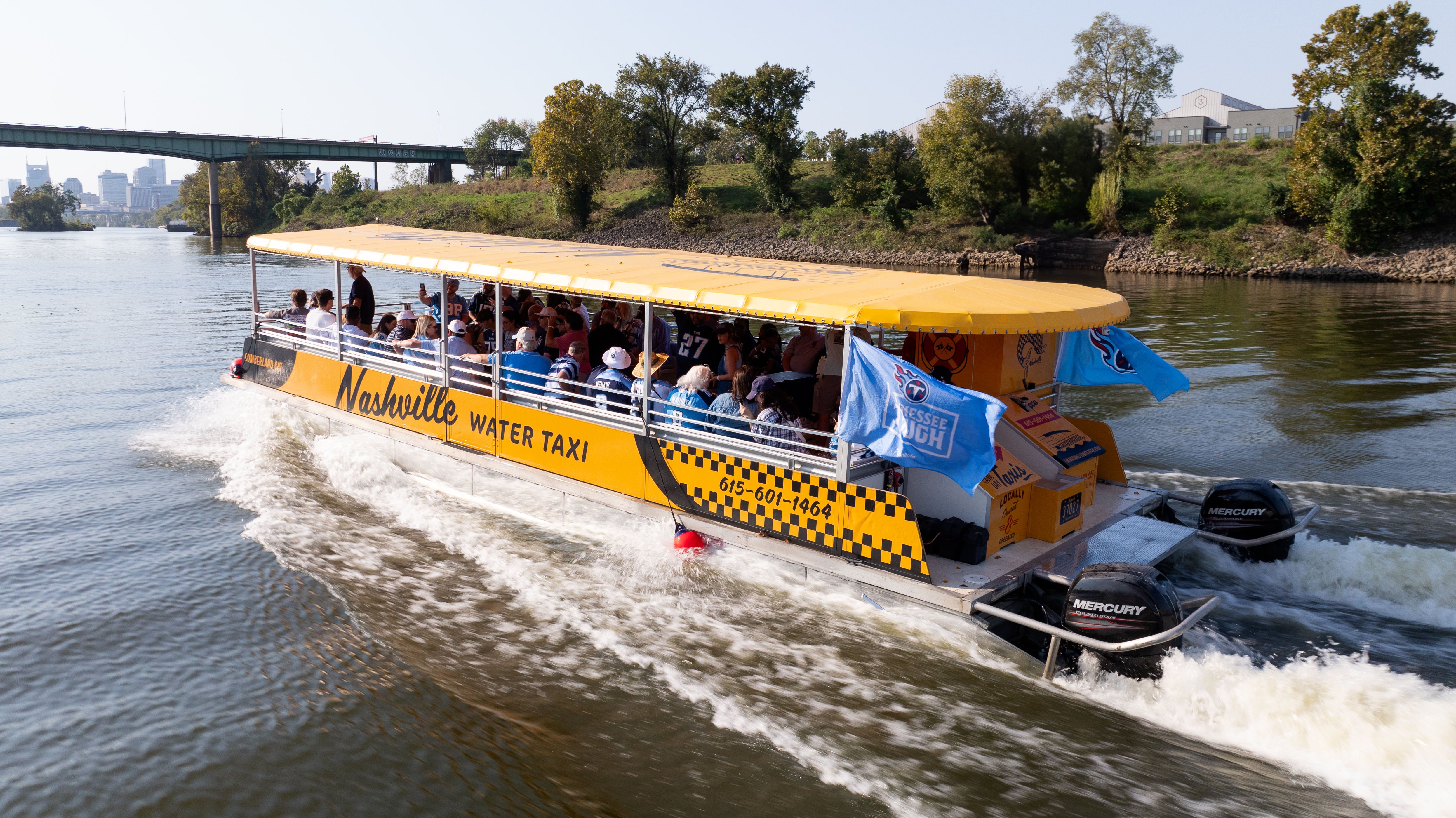 Yellow covered water taxi carrying passengers on the Cumberland River in Nashville, passing under a bridge on a sunny day