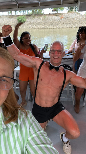 Smiling woman selfie on a riverboat party with a shirtless performer in bow tie and suspenders flexing as friends cheer and sip drinks on the covered deck