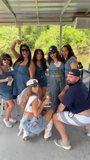 Bachelorette party friends in denim outfits laughing and posing on a covered riverboat deck by green riverside trees