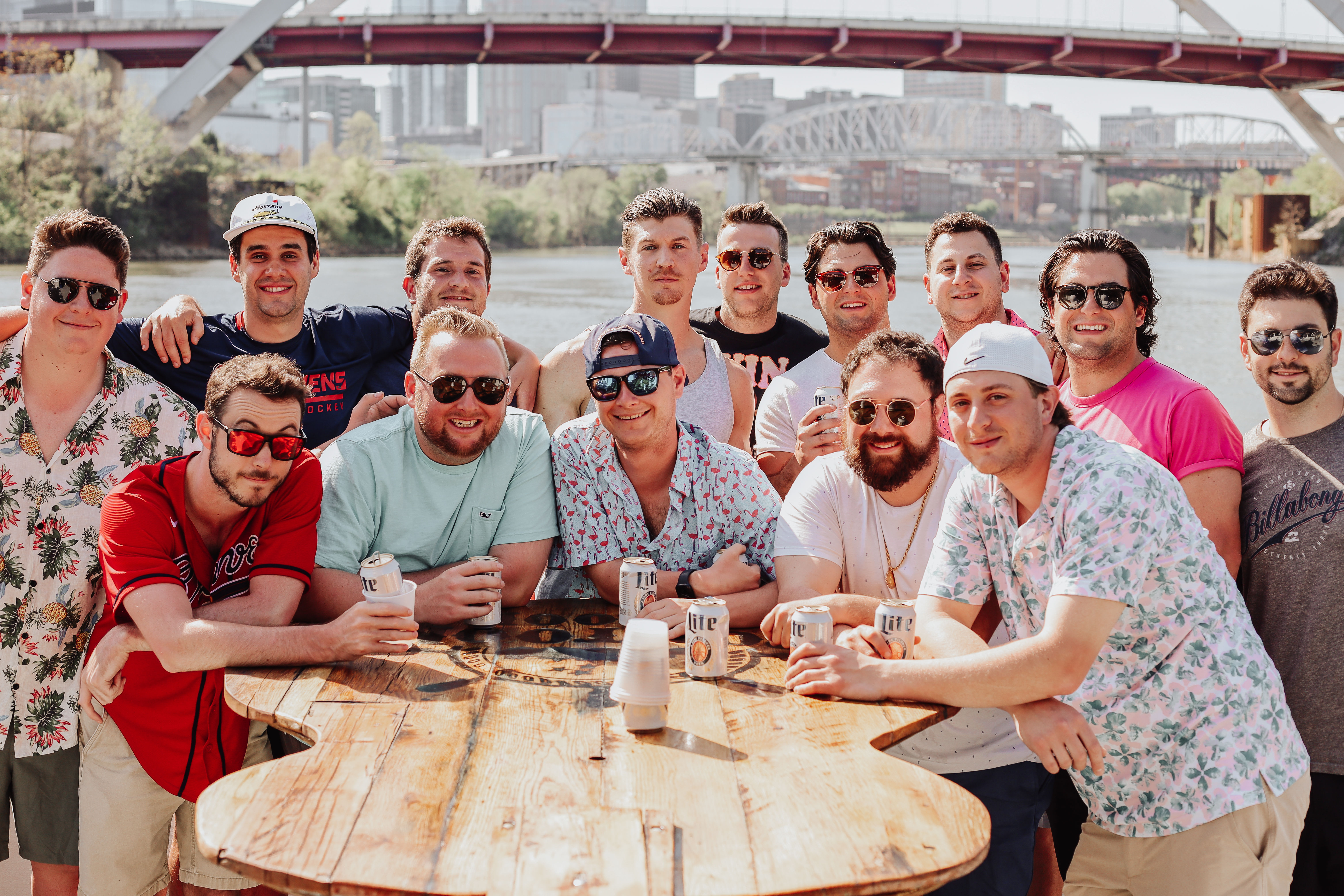 Cheerful group of men in summer shirts gathered around a wooden table by the river, holding canned beers with a city skyline and arched bridge in the background on a sunny day.