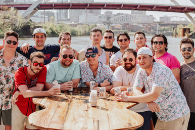 Cheerful group of men in summer shirts gathered around a wooden table by the river, holding canned beers with a city skyline and arched bridge in the background on a sunny day.