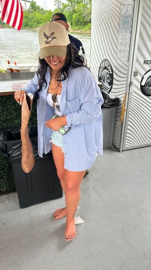 Smiling woman in a tan hat and striped shirt on a riverboat deck, barefoot, dropping a tall brown boot into a trash bin near a restroom door and American flag