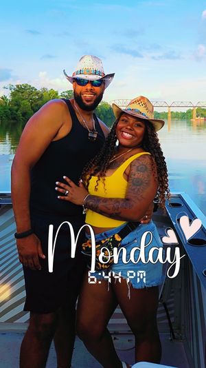Smiling couple in cowboy hats on a boat by a calm river at golden hour, woman in a yellow top and denim shorts and man in a black tank and sunglasses, with a bridge in the background and a 'Monday 6:44 PM' overlay with hearts.