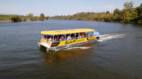 Yellow covered sightseeing riverboat filled with passengers cruising a calm river past tree-lined banks on a sunny autumn day