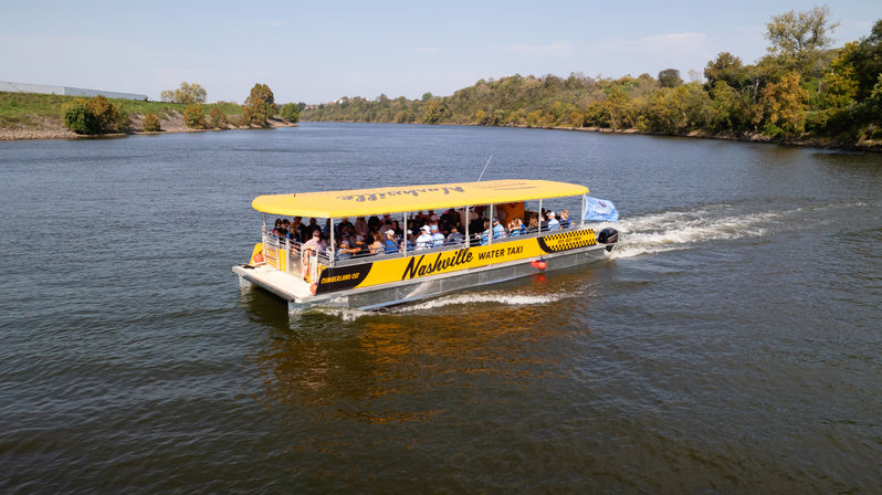 Yellow covered sightseeing riverboat filled with passengers cruising a calm river past tree-lined banks on a sunny autumn day
