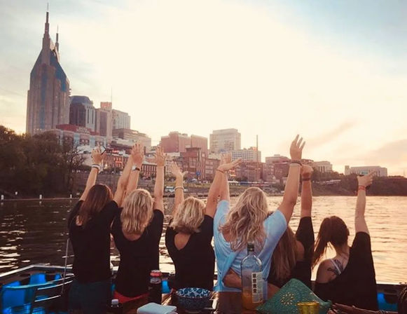Group of friends on a boat raising their hands toward the Nashville skyline at sunset over the river, festive waterfront scene.
