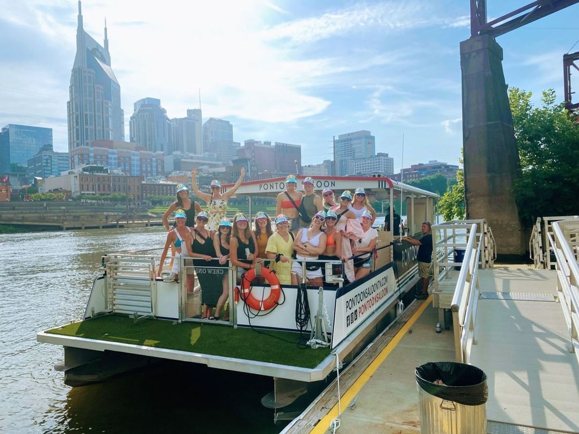 Smiling group of friends on a pontoon boat cruise along a sunny urban riverfront with skyscraper-lined downtown skyline and dock ramp in the foreground.