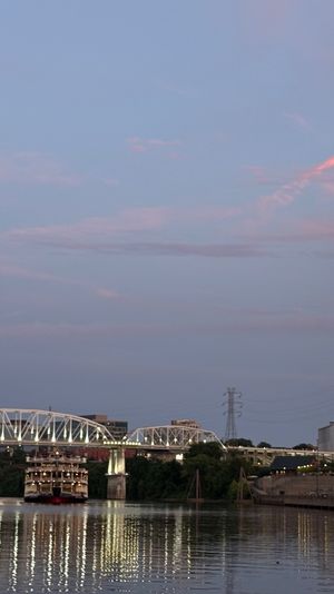 Dusk riverfront skyline with a glass high-rise reflecting pink sunset, illuminated arched bridge, and a paddlewheel riverboat casting shimmering reflections on calm water.