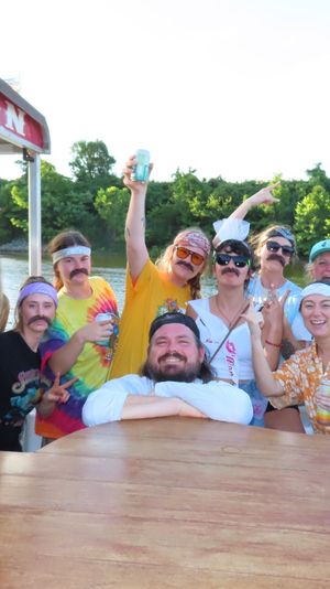 Friends in bandanas and fake mustaches partying on a riverboat saloon deck, holding canned drinks and smiling on a sunny summer river outing