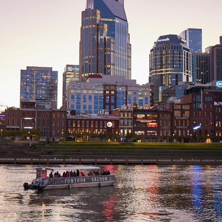 Sunset view of downtown Nashville riverfront with illuminated skyscrapers and historic brick buildings, and a lively pontoon tour boat casting colorful reflections on the water.