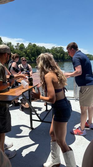 Summer pontoon party on a river: people gathered around a wooden table playing a drinking game, trees along the shoreline and a rail bridge in the distance.