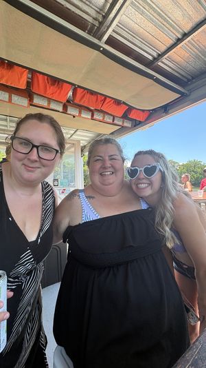 Three smiling women on a sunny outdoor deck, close-up group selfie — left woman in black-and-white dress and glasses holding a can, center woman in a black off-shoulder dress, right woman wearing heart-shaped sunglasses and a bikini top, with other patrons and trees visible in the background.