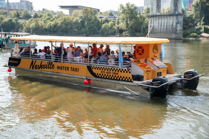 Yellow covered water taxi crowded with passengers cruising a river in Nashville, Tennessee, with trees and city buildings along the riverbank.