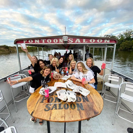 Group of smiling women on a covered pontoon party boat cruising a river, raising drinks around a wooden table for a bachelorette celebration under a cloudy evening sky