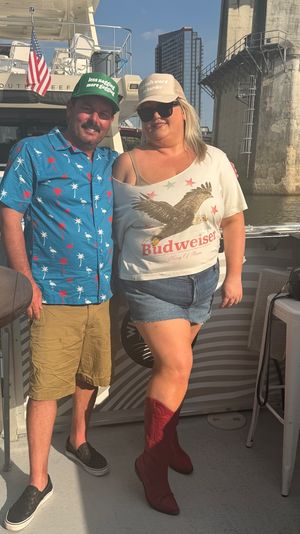 Two smiling adults on a leisure boat by an urban bridge and river at golden hour, man in a tropical shirt and cap, woman in a vintage beer T-shirt, denim shorts and red boots, American flag visible.