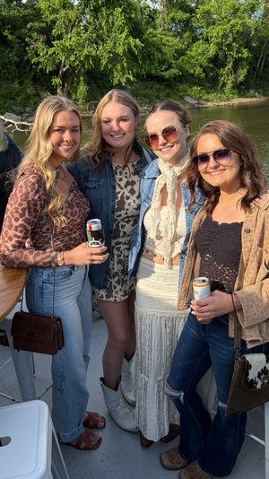 Four smiling women posing on a riverboat deck by a tree-lined riverbank, wearing casual summer outfits and sunglasses and holding canned drinks — outdoor riverside gathering.