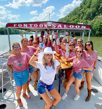 Bachelorette party of women on a pontoon saloon boat cruising a sunny river, all smiling and raising drinks; bride-to-be in a white hat and veil, casual summer denim and boots, wooded riverbanks and blue sky