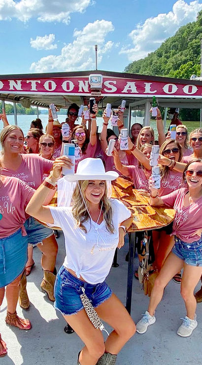 Bachelorette party of women on a pontoon saloon boat cruising a sunny river, all smiling and raising drinks; bride-to-be in a white hat and veil, casual summer denim and boots, wooded riverbanks and blue sky