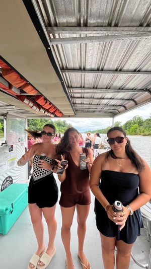 Three young women in summer outfits and sunglasses holding canned drinks on a covered pontoon boat cruising a tree-lined river, with orange life jackets overhead and fellow passengers in the background.
