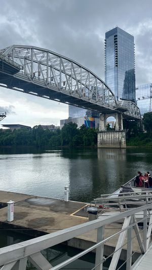 Steel truss bridge arching over a calm river with mirrored glass skyscrapers and a colorful mural on the far bank, people boarding a boat at a riverside dock under a cloudy sky.