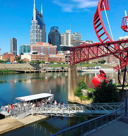 Downtown Nashville skyline along the Cumberland River with a docked tour boat, bright red industrial bridge sculpture and shimmering reflections on a sunny day.