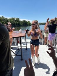 Woman in heart-shaped sunglasses, purple bikini top, denim shorts and white cowboy boots dancing with drinks on a sunny riverboat deck, tree-lined river and bridge in the background — summer boat party.