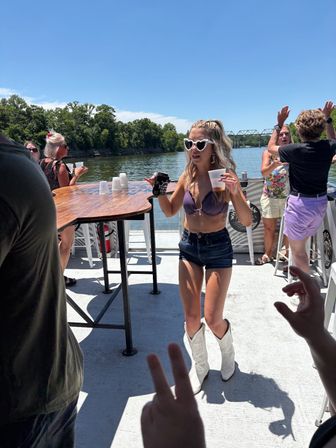 Woman in heart-shaped sunglasses, purple bikini top, denim shorts and white cowboy boots dancing with drinks on a sunny riverboat deck, tree-lined river and bridge in the background — summer boat party.