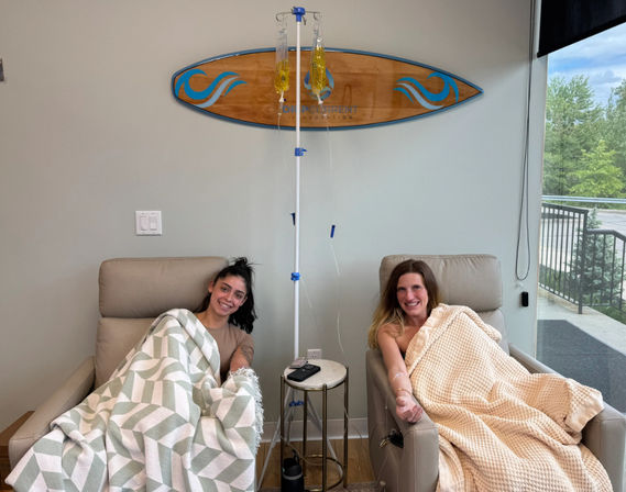 Two women reclining in infusion lounge chairs receiving IV vitamin drips, cozy blankets, surfboard wall decor and a large window showing outdoor greenery